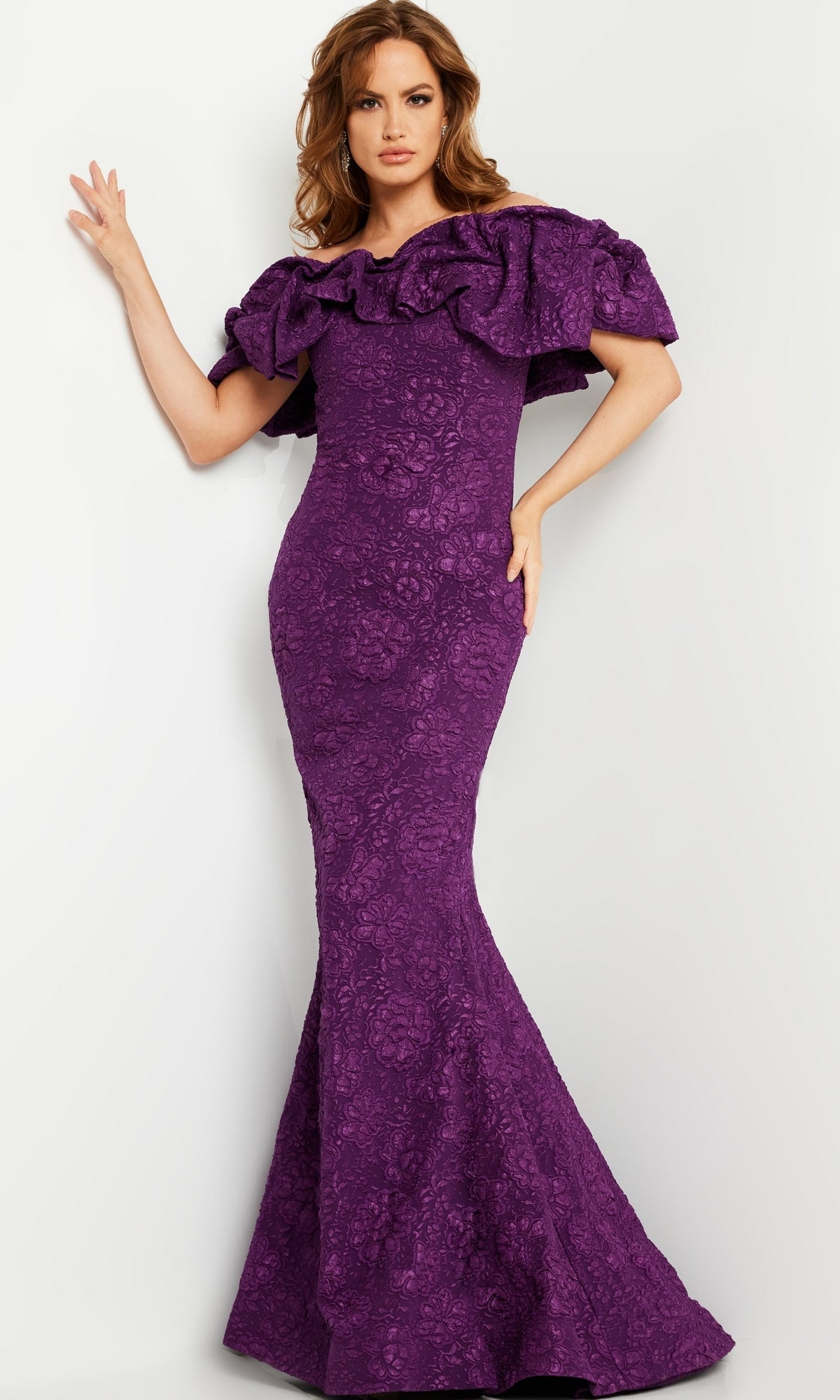 A woman poses elegantly in the Jovani Long Brocade Mermaid Prom Dress 23847, featuring a fitted silhouette, floor-length skirt, ruffled off-the-shoulder neckline, and floral texture against a plain background.